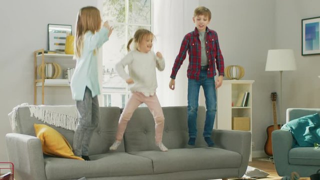 Two Cute Little Girls and Young Adorable Boy Have Fun, Jumping High on a Couch at Home. Happy Children Playing in the Sunny Living Room. In Slow Motion.