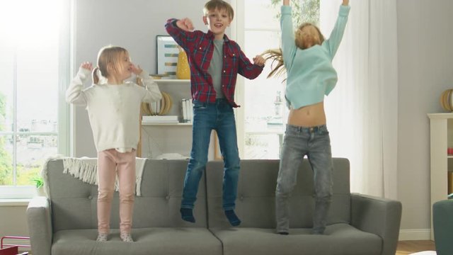 Two Cute Little Girls and Young Adorable Boy Have Fun, Jumping High on a Couch at Home. Happy Kids Dancing on a Sofa in the Sunny Living Room. In Slow Motion.