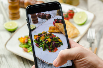 Young man taking photo of fish and vegetables plate on smartphone. Taking food photo with mobile...