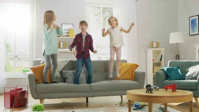 Two Cute Little Girls and Young Adorable Boy Have Fun, Jumping High on a Couch at Home. Happy Kids Dancing on a Sofa in the Sunny Living Room. 