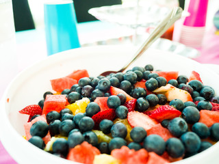 Close up photograph of a home made fruit salad for a gender reveal party containing blueberries, strawberries, pineapple, and watermelon in a white serving bowl and blue and pink decor beyond.