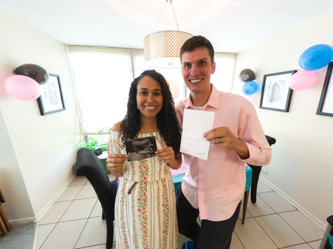 A Mixed Caucasian And African American Biracial Couple Dressed Up In A Pink Shirt And Dress Smile And Hold Up Card And Ultrasound Revealing They Are Expecting A Boy With Decorations In The Background.