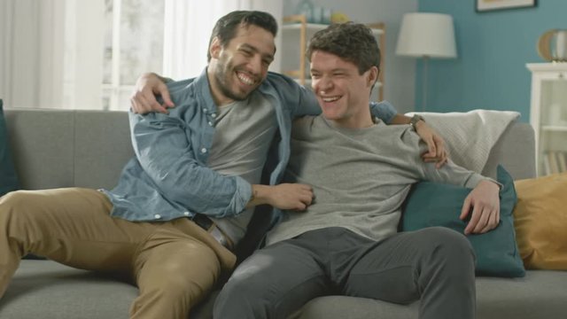 Sweet Male Gay Couple Full Around on a Sofa at Home. Boyfriend Runs and Jumps into Hands of His Partner. They Hug. They are Happy and Laughing. They are Casually Dressed and Room Has Modern Interior.