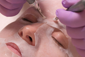 Fototapeta premium Close-up of electrodes in the hands of a beautician, touching the female cheek through a mask. Woman receiving stimulating facial treatment with microcurrent equipment. Wrinkle smoothing.
