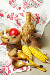 Still life with a wicker bottle, corn, tomato, garlic and a woven towel.