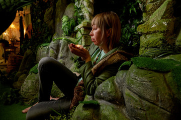 Girl in a green jacket with blond hair with a small candle in her hands near an artificial rock with a grotto