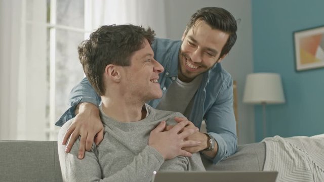 Sweet Male Gay Couple Spend Time at Home. Young Man Works on a Laptop, His Partner Comes From Behind and Gently Embraces Him. They Laugh and Touch Hands. Room Has Modern Interior.
