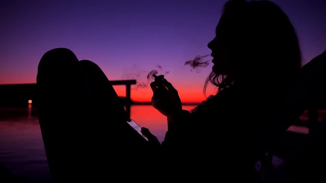 Silhouette of Young Unidentified Woman Smoking Weed Joint or Cigatette Againts Amazing Beach Sunset near Water. 4K Slowmotion.