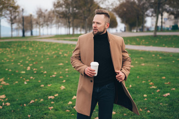 Handsome bearded young man in sunglasses drinking coffee in park, copy space on cup