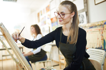 Beautiful fair-haired girl in glasses dressed in black blouse and an apron sits at the easel and paints a picture in the art studio
