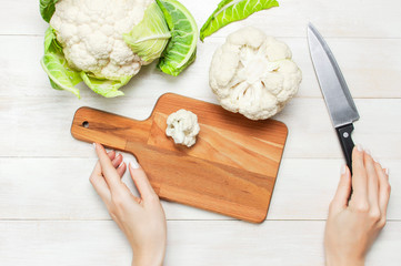 Female hands cut cauliflower with knife on cutting board, kitchen towel on rustic white wooden...
