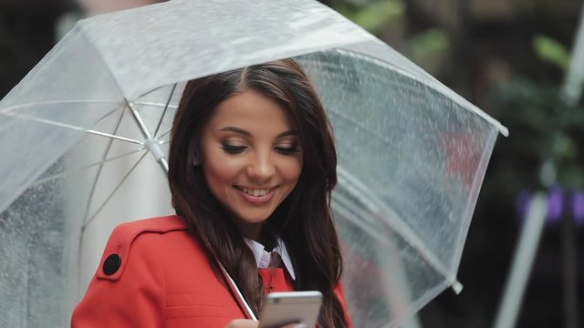 Beautiful young business woman using smartphone on the street in rainy weather, smiling, holding umbrella, communication concept