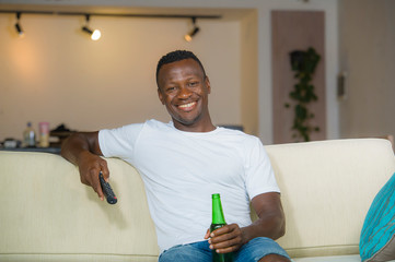 young attractive and happy black afro American man holding TV remote controller watching television movie laughing relaxed drinking beer in living room couch at home