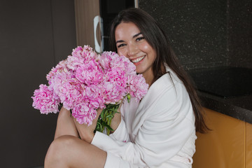 Portrait of a young beautiful brunette in a white robe and a bouquet of pink peonies at home. A girl with a bouquet of flowers.