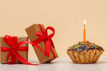 Tasty birthday muffin with burning festive candle and gift boxes on beige background.