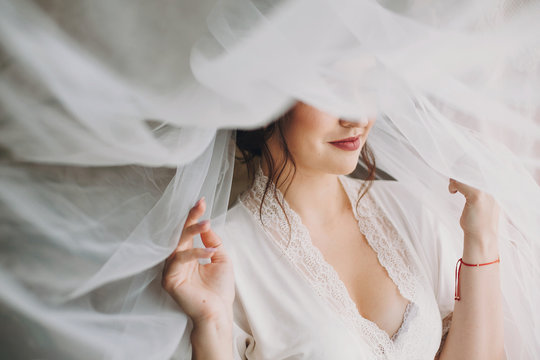 Beautiful stylish brunette bride posing in silk robe under veil in the morning. Happy woman model with perfect makeup and hairstyle, getting ready for wedding day