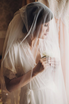 Beautiful stylish brunette bride posing in silk robe under veil in the morning. Sensual portrait of happy woman model with boutonniere in hands getting ready for wedding day