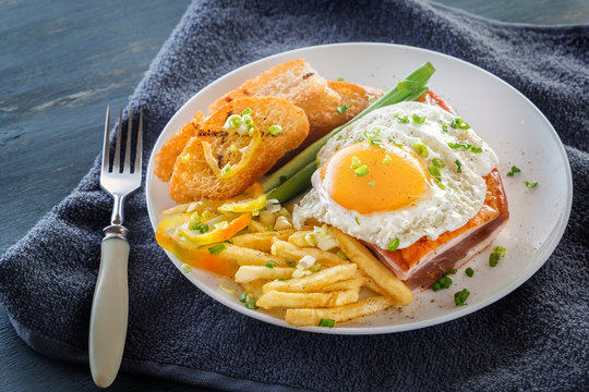 Fried egg with bacon in a plate with fried pieces of bread, greens and french fries on a gray wooden table. Close-up