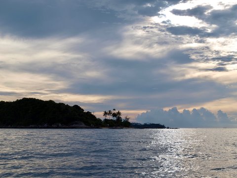 Silhouette of tropical island with palms in the sea in sunset. Panoramic view from water. A perfect picture of a tropical seaside resort and boat trip.  Amazing seascape in sundown evening time. Cloud