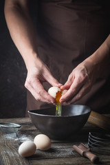 Professional chef hands are breaking an egg into bowl to make dough on wooden table, over dark background