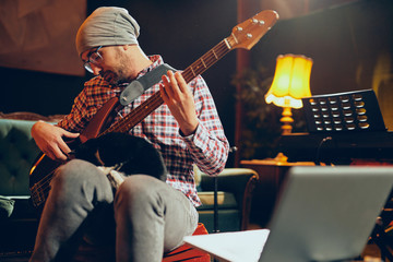 Obraz premium Young Caucasian man with hat on head playing bass guitar while sitting in home studio.