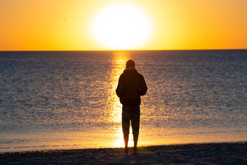 Fototapeta premium Man stands on the beach next to surf, admires amazing sunset