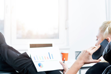 Rear view of a blonde girl checking sales chart in the work or home office while talking on a mobile and relaxing her with feet on the desk.