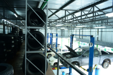 the blur of grey pick up car and other on the lift inside the car service center with the many tyres on shelf at above and blue oil tank at bottom with bright daylight from outside