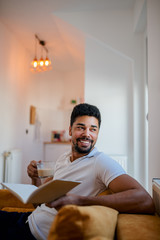Fototapeta premium Handsome young man sitting on sofa, reading a book and drinking coffee.