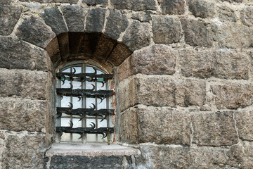 Window with wrought iron grille on dark stone wall. Wall of old house. Building facade.
