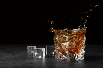 Splashing golden whiskey in glass with ice cubes on table. Space for text