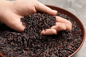 Gordijnen Woman holding grains near plate with black rice on table, closeup © New Africa