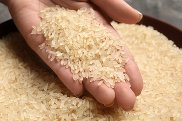 Woman holding grains near plate with parboiled rice on table, closeup