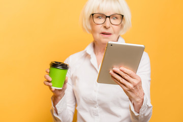 Happy senior woman using tablet while drinking coffee isolated over yellow background.