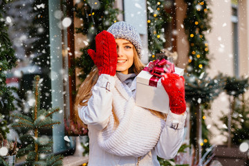 Good winter holiday mood. Excited blond woman closing on eye with hand in red mittens and holds gift box with red bow in other hand against shop window with lights and decorations in winter day. Snow