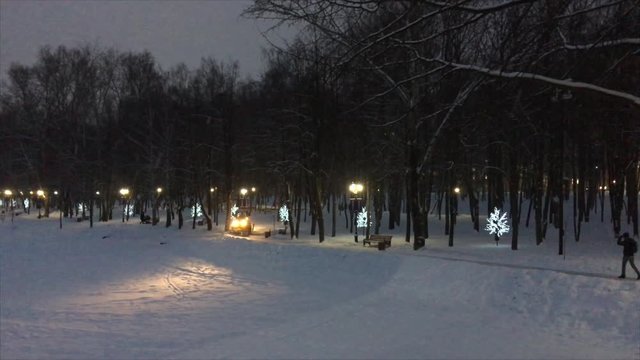 A small snow-removing tractor with headlights turned on cleans the footpath in the city park under the falling snow and in the evening lights.