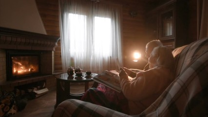 An elderly couple considers a family photo album wrapped in a blanket sitting by a cozy fireplace. Golden wedding, happy marriage, grow old together - Powered by Adobe