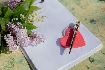 Mock up letter about love.Red wooden heart lies on blank white paper on a wooden table with spring flowers, a greeting card for Valentine's Day with a place for your text. Flat lay, lilac and lilies