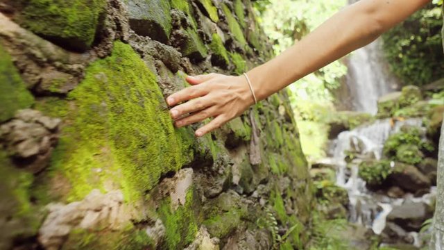 Young Woman in White Dress Walking in Tropical Jungle to Beautiful Waterfall and Touching with Hand Rock Wall with Moss. Calm and Carefree Lifestyle Travel 4K Slowmotion Footage. Bali, Indonesia.