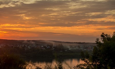 Fototapeta na wymiar Dawn over the river. Sunrise over the village. Morning rays of the sun. The first rays of the rising sun. The village is at dawn. Smoke from the pipes of houses. Summer sky in the morning. The village
