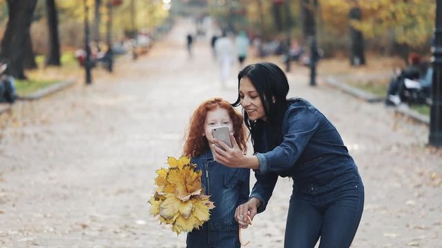 A beautiful mom and her cute little daughter walking in the autumn park. They making a selfie on a smartphone, laughing and have fun