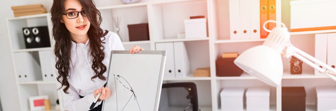 A beautiful young girl stands near an office desk and draws a magnetic marker on the magnetic board.