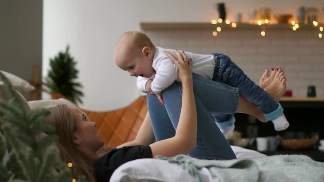 Mom plays with Baby in white tank top lying on the bed, baby flying and laughing. Time lapse of playing together mother and child. Flight simulation
