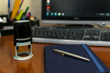 Stamp for documents, notepad and pen lie on a wooden office desk on a computer and stationery background, closeup
