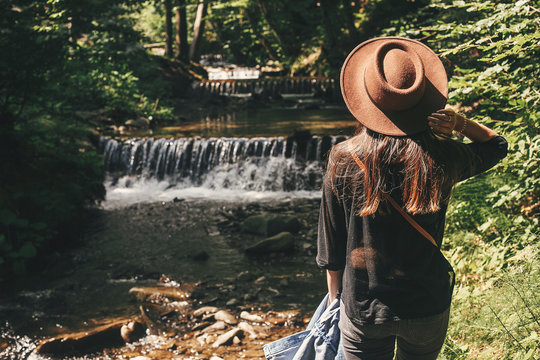Stylish hipster girl traveling in woods, back view. Young woman in hat exploring in summer forest, looking at river. Travel and wanderlust concept. Space for text