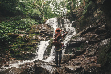 Stylish hipster girl in hat traveling in mountains. Young woman with backpack exploring in summer forest, looking at waterfall. Travel and wanderlust concept. Space for text
