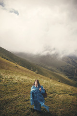 Stylish hipster girl in blue raincoat and with backpack standing on top of misty mountains and clouds. Young woman traveler exploring mountains. Travel and wanderlust concept. Atmospheric moment