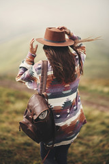 Stylish hipster girl in hat walking on top of mountains. Happy young woman with backpack exploring misty mountains. Travel and wanderlust concept. Amazing atmospheric moment