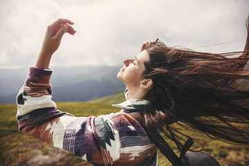 Stylish hipster girl with backpack  and windy hair on top of mountains. Portrait of happy young woman relaxing. Carefree mood. Amazing atmospheric moment