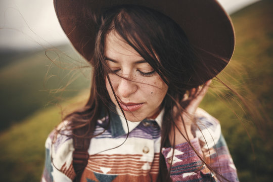 Portrait of happy calm young woman with windy hair. Carefree mood. Amazing atmospheric moment. Stylish hipster girl in hat and backpack walking on top of mountains